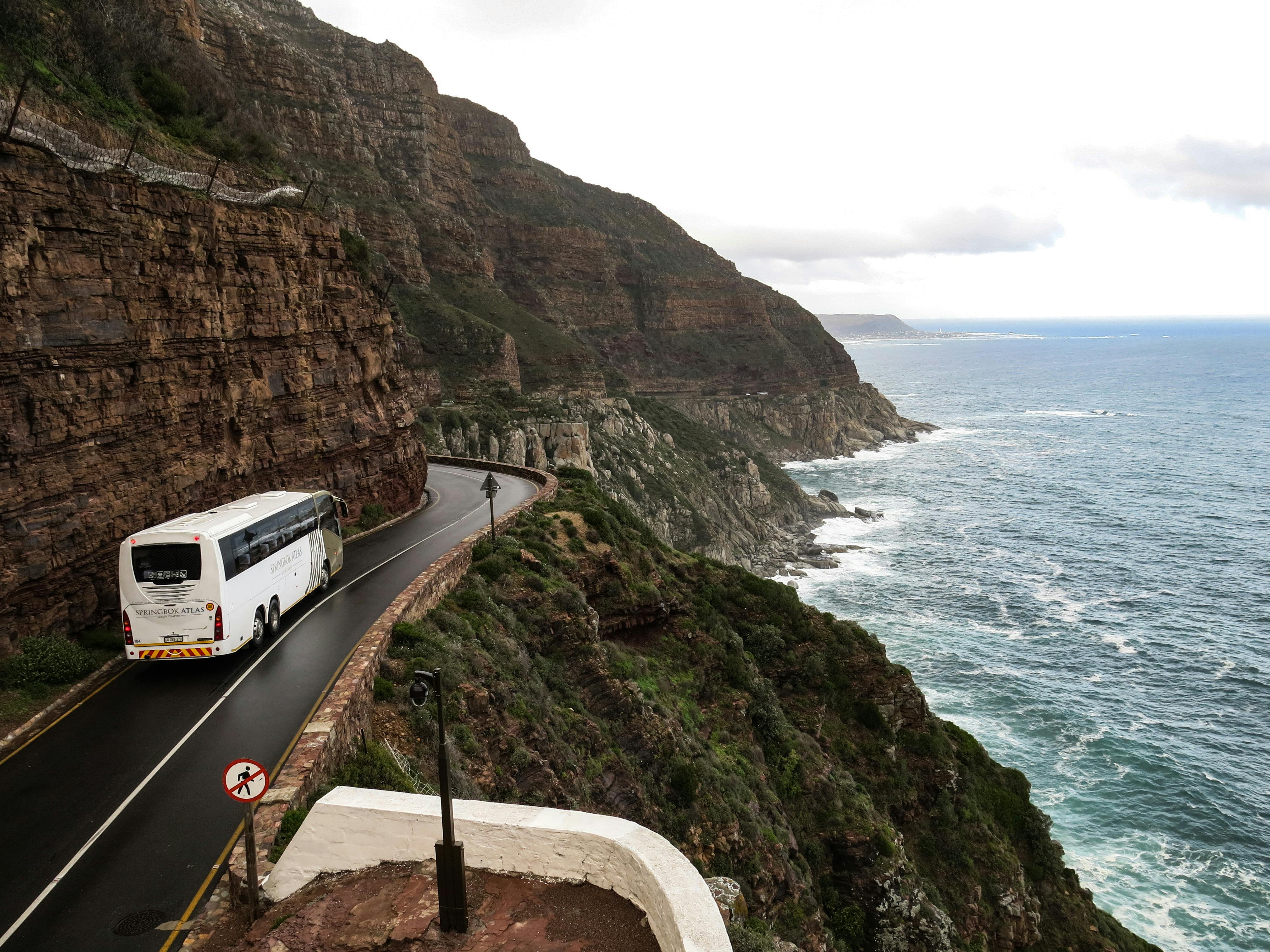 Photo of a white bus driving along a cliffside road above a rocky beach